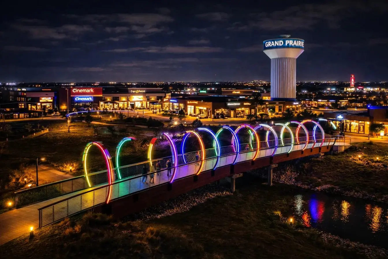 Rainbow-lit pedestrian bridge at night in Grand Prairie Texas with water tower and retail centers in the background